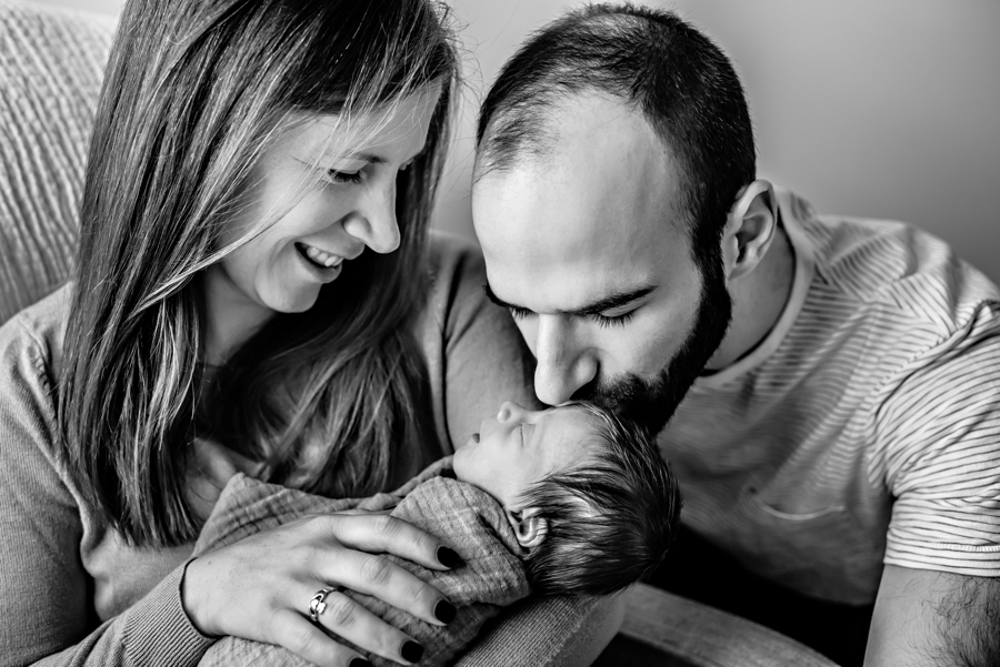 A new father kisses his newborn baby girl in the arms of her mother.